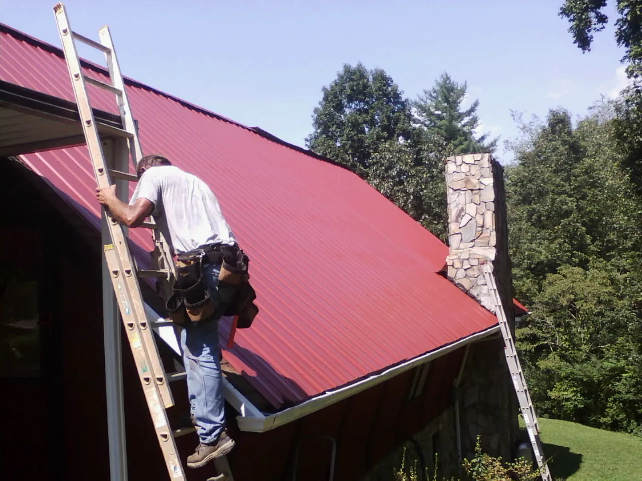 Roofer on a ladder beside a red metal roof and stone chimney — Soffit & Fascia Repair by Roofing Brothers in Pleasant Valley